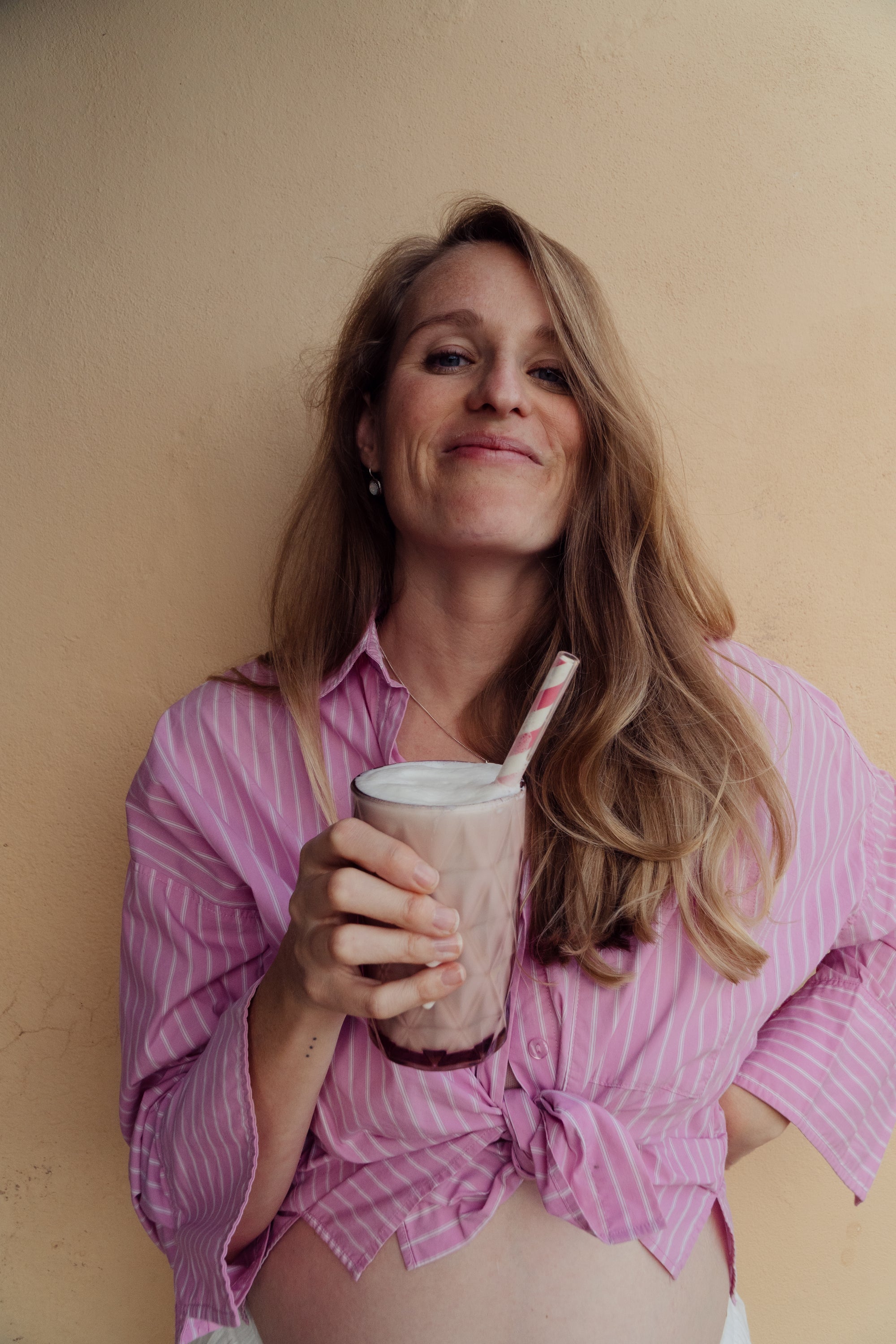 Woman in a pink striped shirt holding a pink cup filled with the MomShake with a straw against a beige wall on Ibiza