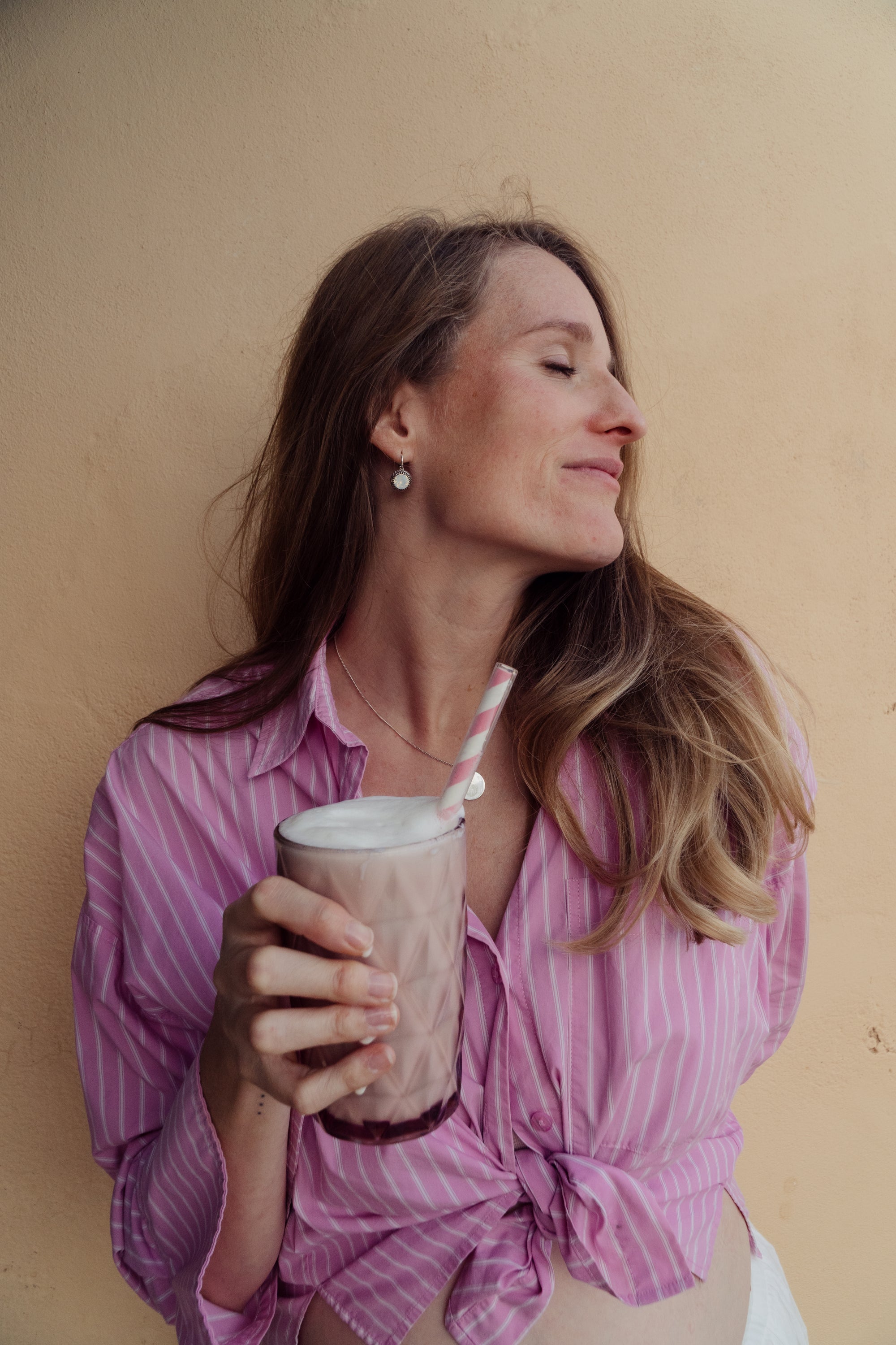 Woman in a pink striped shirt holding the MomShake in a pink glass with a straw against a beige wall on Ibiza